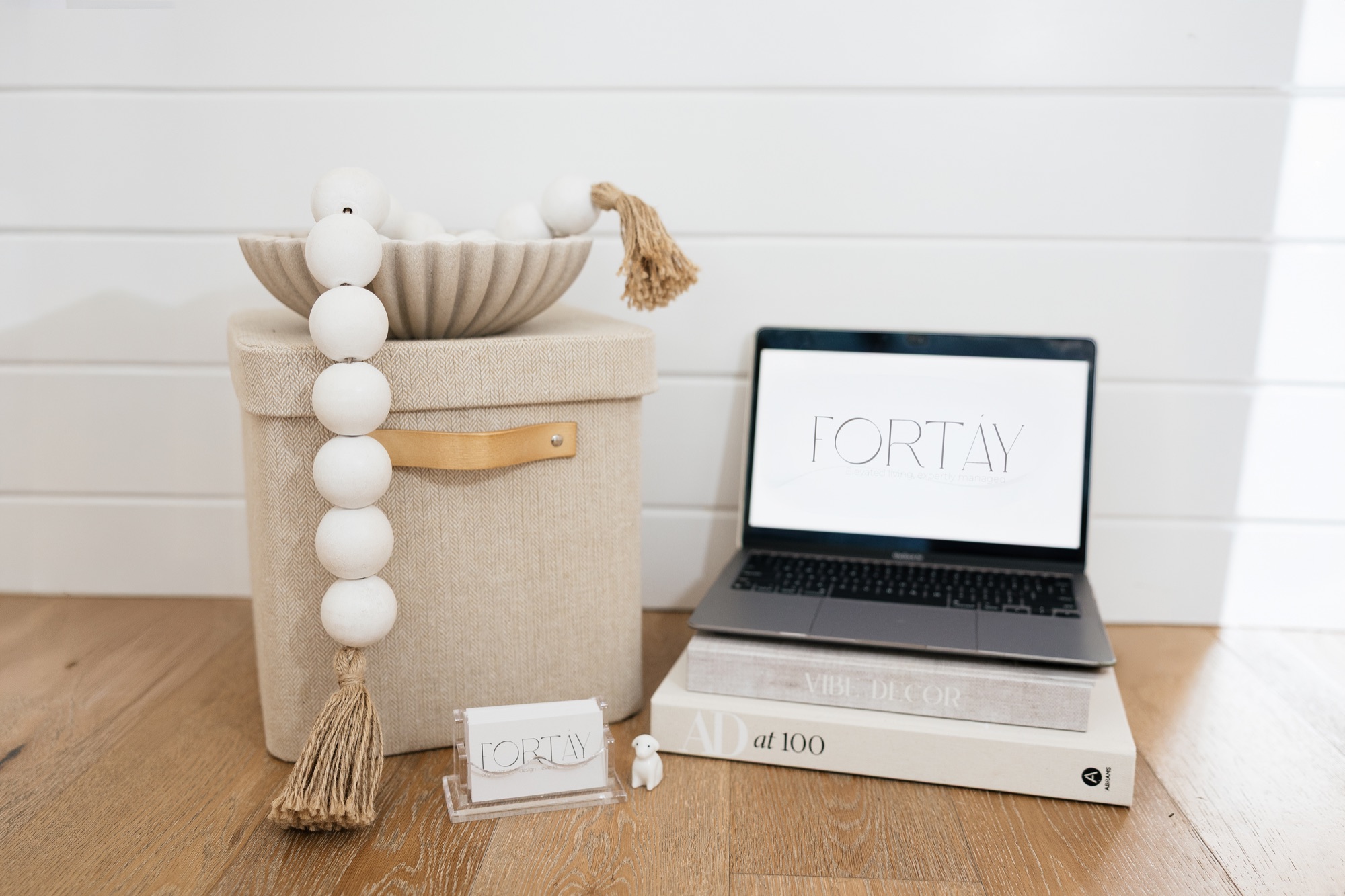 Laptop displaying the Fortáy logo resting on a stack of design books beside a woven basket and wooden bead garland